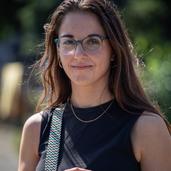 Jana Odvárková 在克拉德诺摆姿势拍照。她戴着眼镜，穿着黑色无袖上衣，戴着项链，肩上挂着一条带图案的肩带。背景被绿色植物模糊了